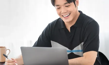 A student smiling at a laptop while taking notes at a desk with books and a coffee mug nearby.