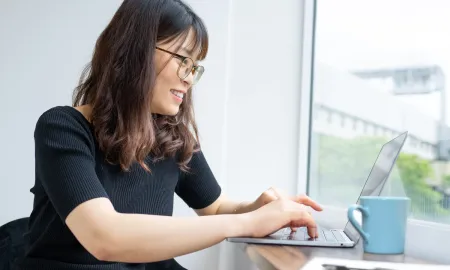 A woman in glasses smiling while typing on her laptop during an online event, with a coffee mug nearby.