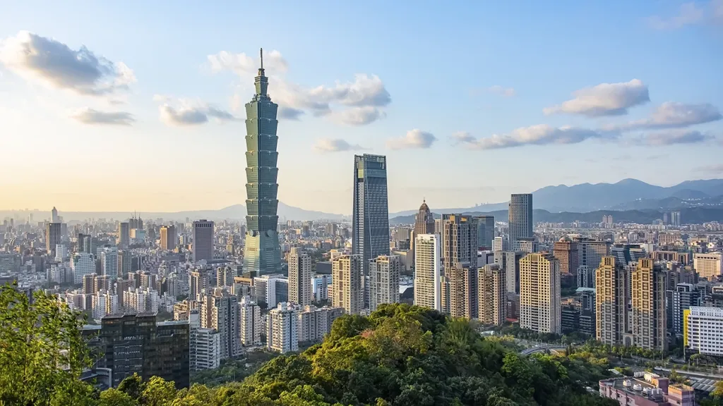Skyline of Taipei featuring the Taipei 101 tower, viewed from a green hillside with mountains in the background.